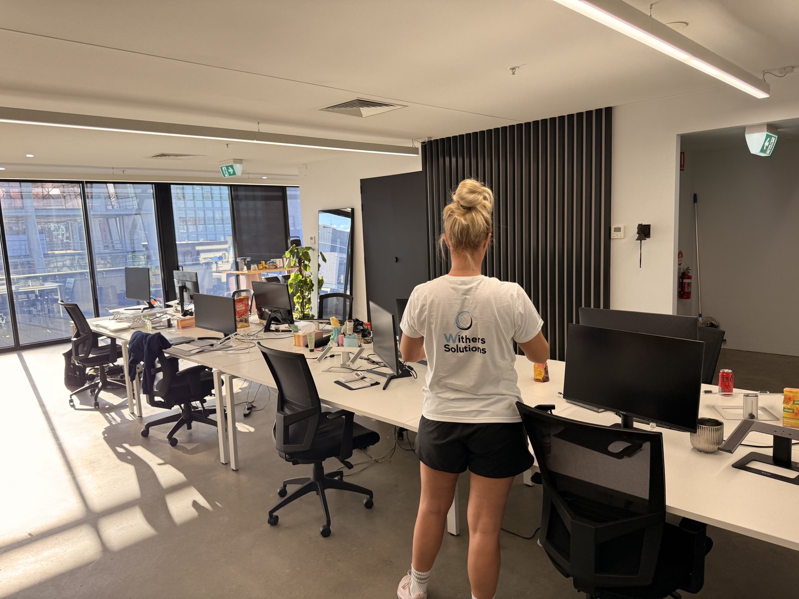 A cleaner in a Withers Solutions T-shirt wiping down long white workbenches in a sunlit open-plan office in Richmond