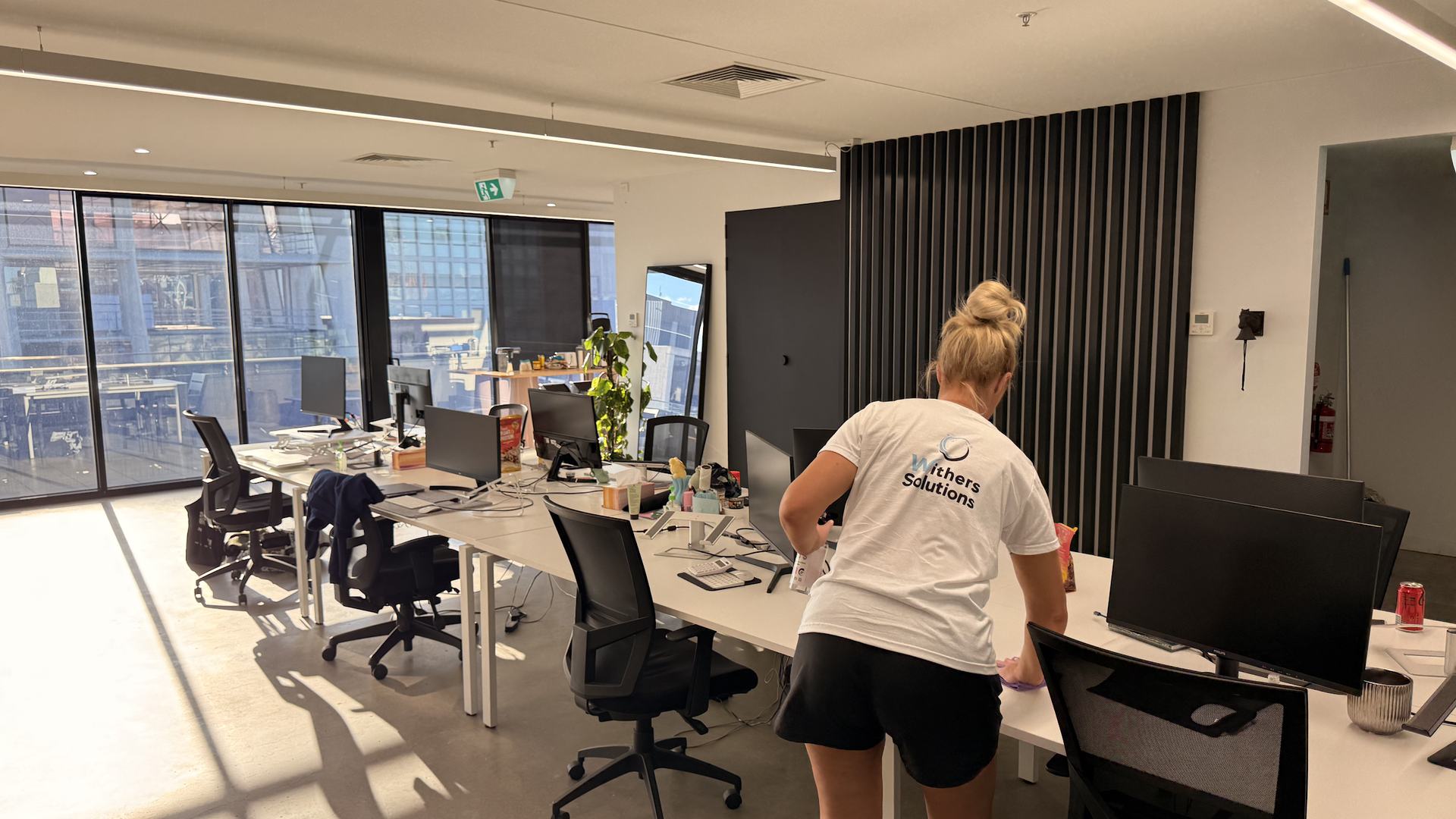 A Withers Solutions cleaner wiping down desks in an open-plan office with floor-to-ceiling windows